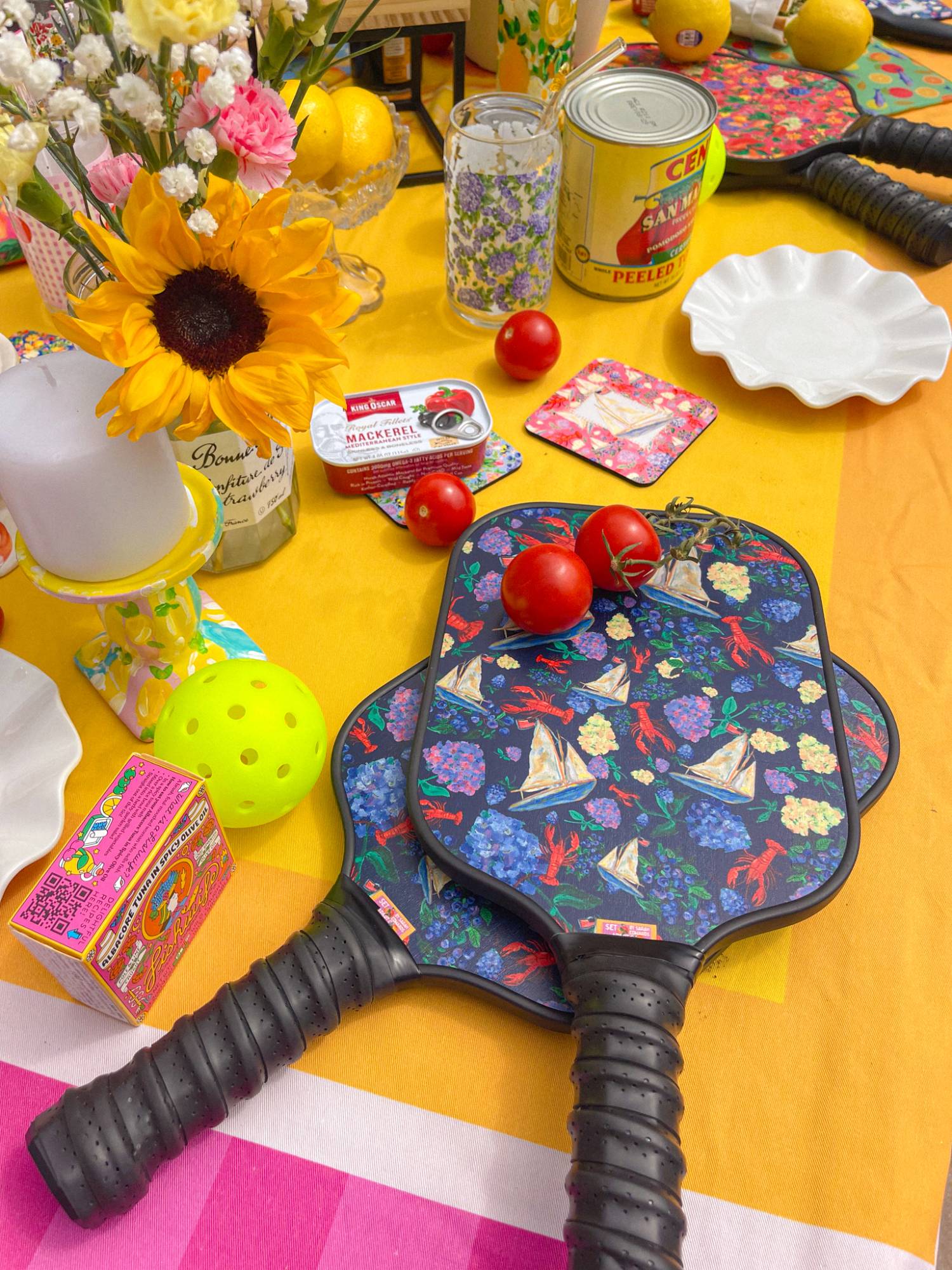 Navy sailboat and lobster pickleball paddles on a sunny yellow table with sunflowers and tomatoes