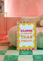 Reading Is Cheaper Than Therapy 24x36 bookish wall art poster propped beside a woven pouf on a green checkered floor