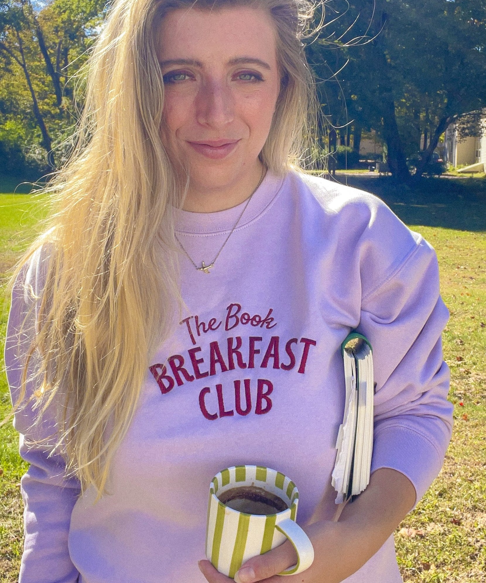 Woman wearing the lavender Book Breakfast Club embroidered sweatshirt holding a mug and book
