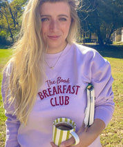 Woman wearing the lavender Book Breakfast Club embroidered sweatshirt holding a mug and book