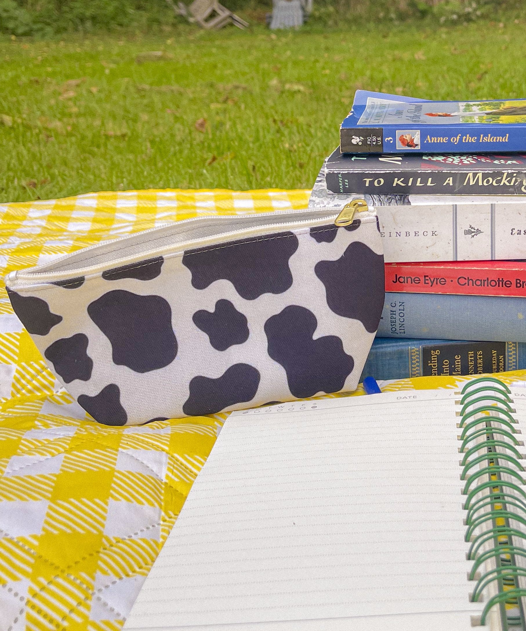 Cow print accessory pouch with books and a notebook on a yellow gingham blanket