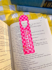 Pink checkered Reading Is Cheaper Than Therapy metal bookmark clipped in an open book