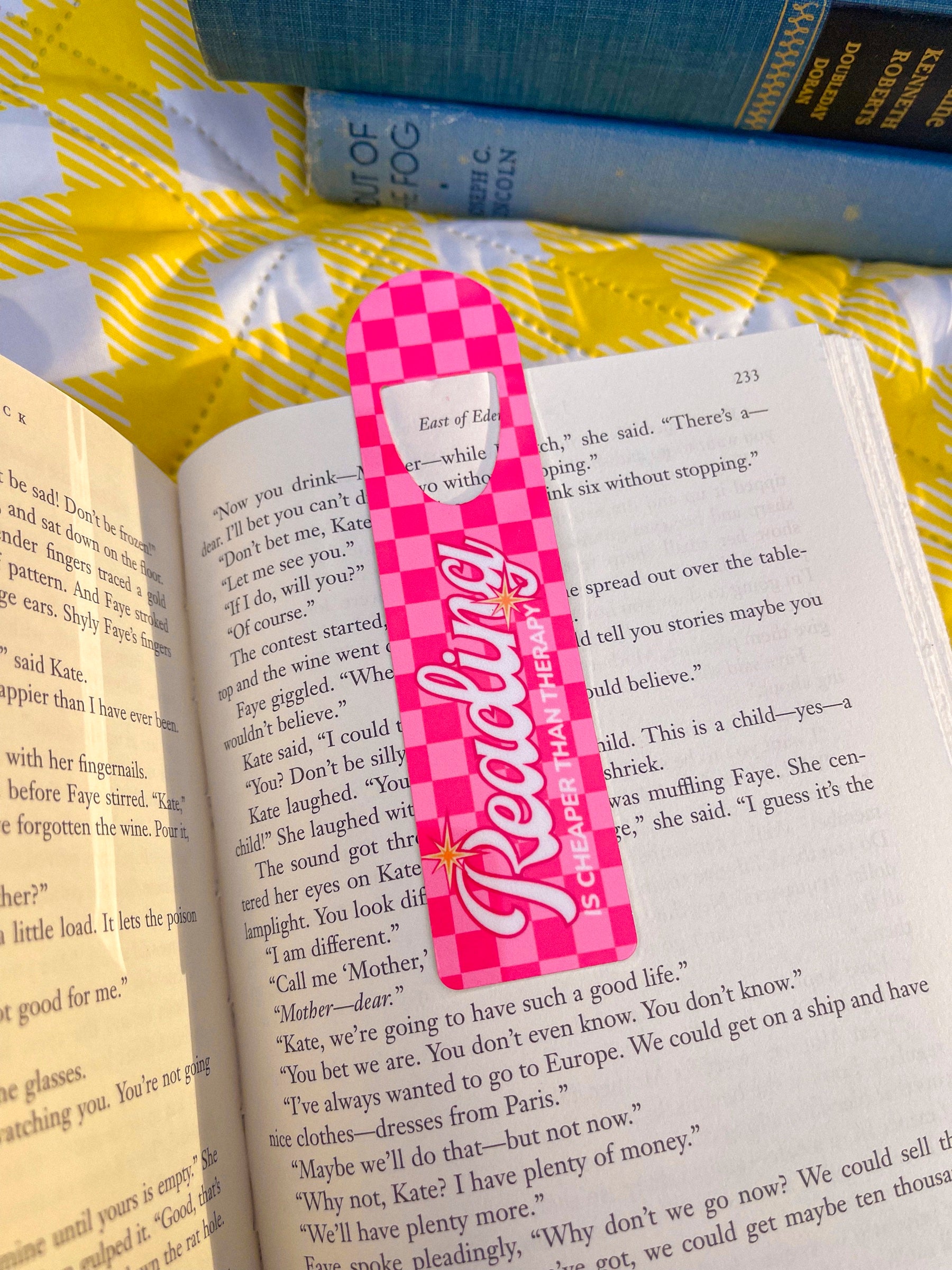 Pink checkered Reading Is Cheaper Than Therapy metal bookmark clipped in an open book