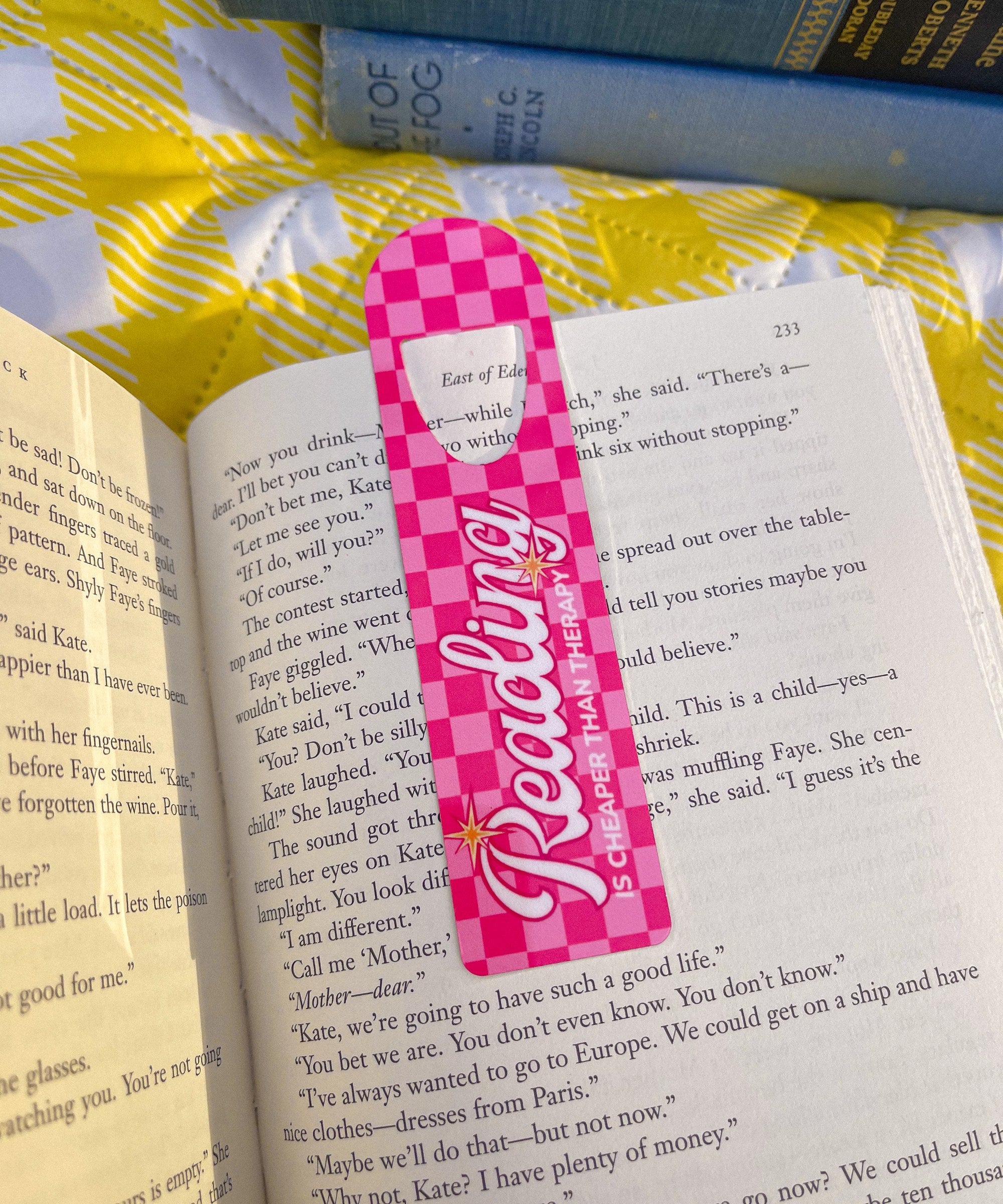 Pink checkered Reading Is Cheaper Than Therapy metal bookmark clipped in an open book