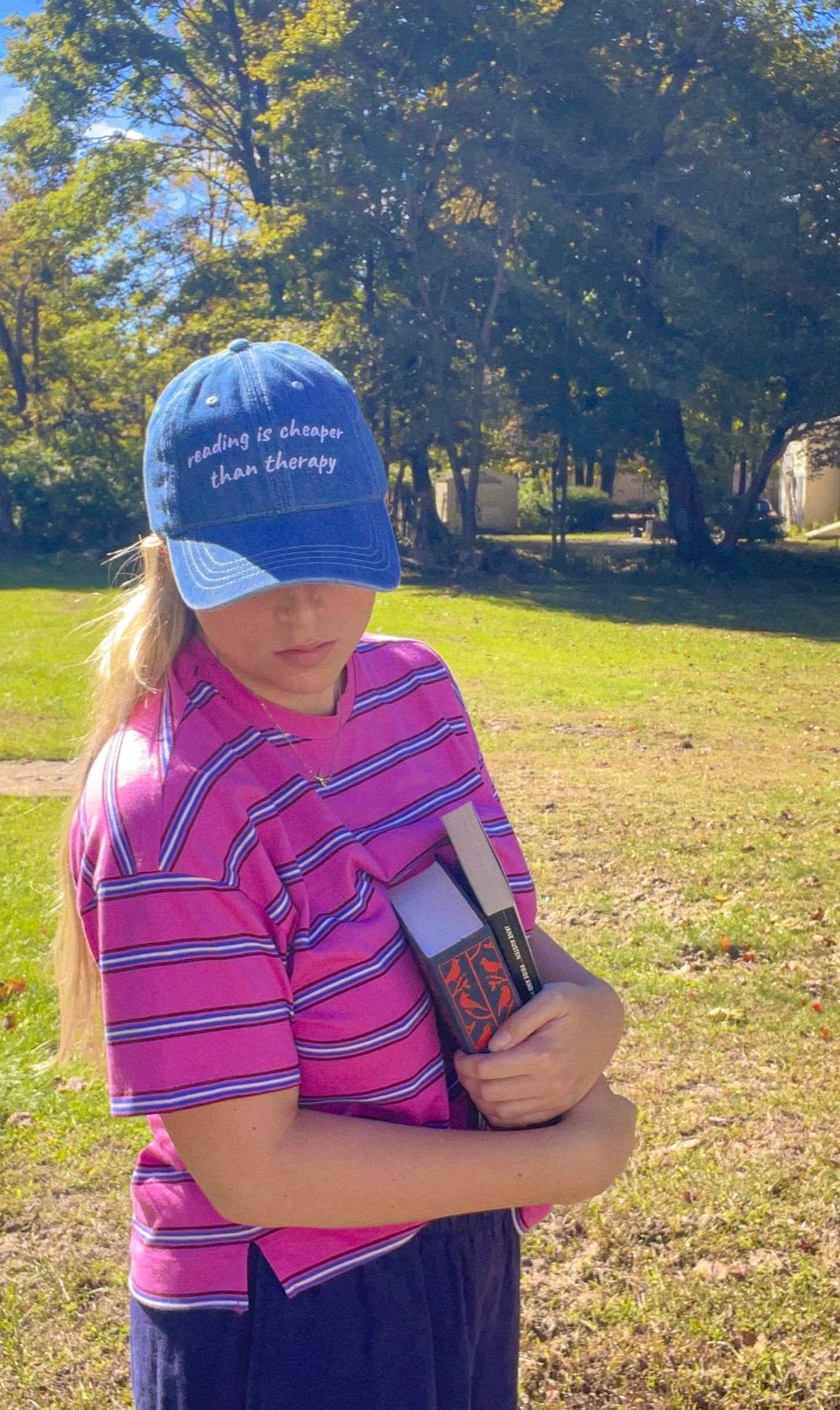 Woman wearing the Reading Is Cheaper Than Therapy embroidered denim hat holding a book