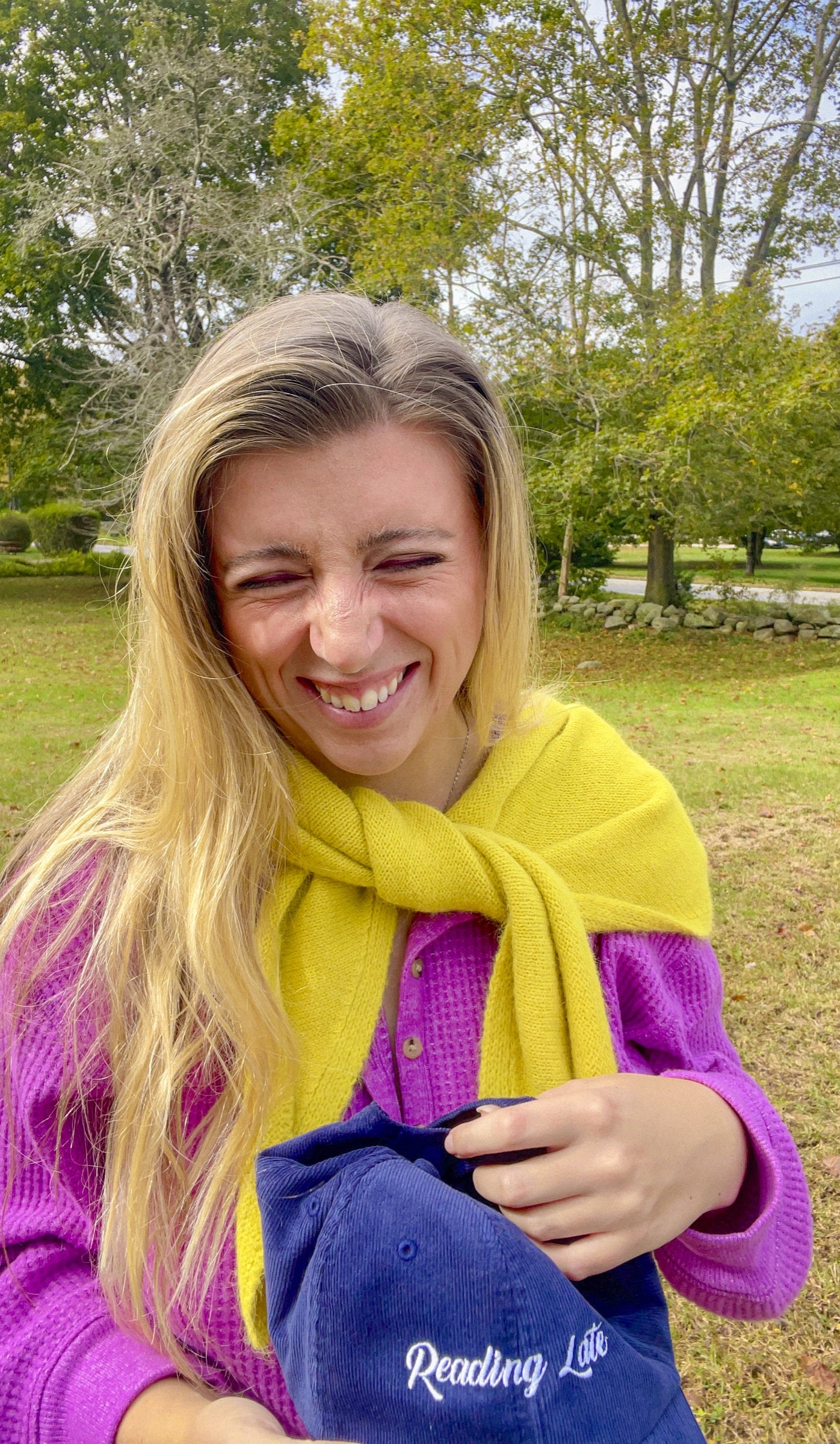 Sarah laughing in a purple cardigan and yellow scarf, holding a Reading Late corduroy hat