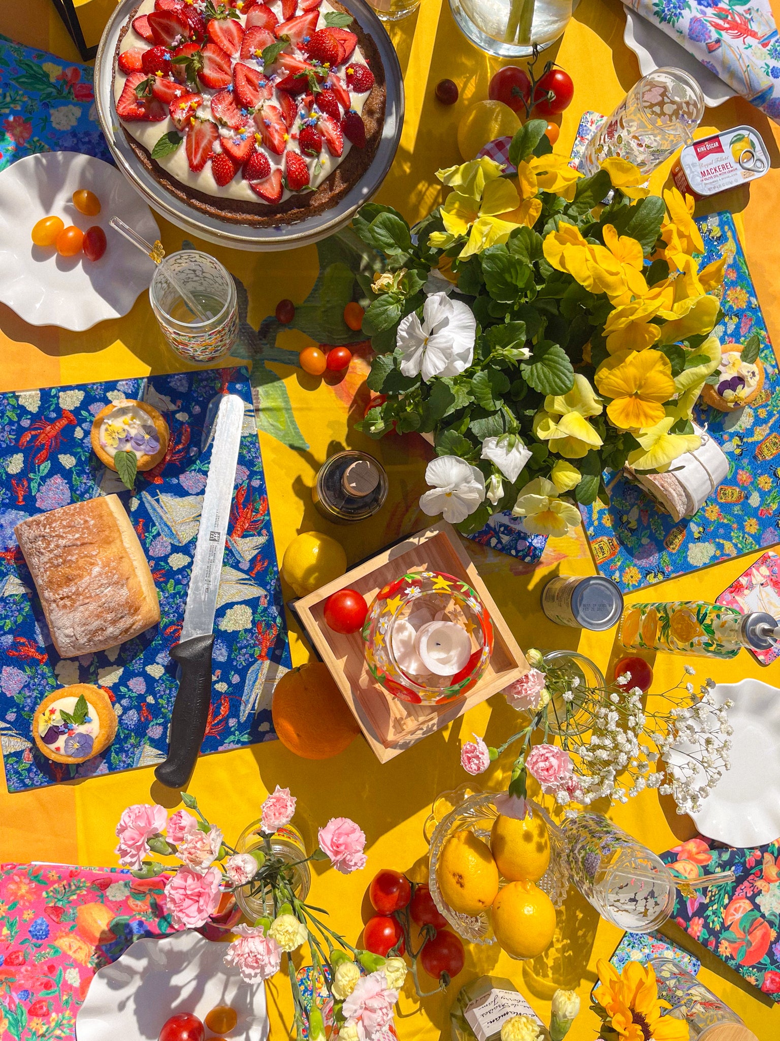 Overhead table with a strawberry cake, painted glass cutting boards, pansies and lemons